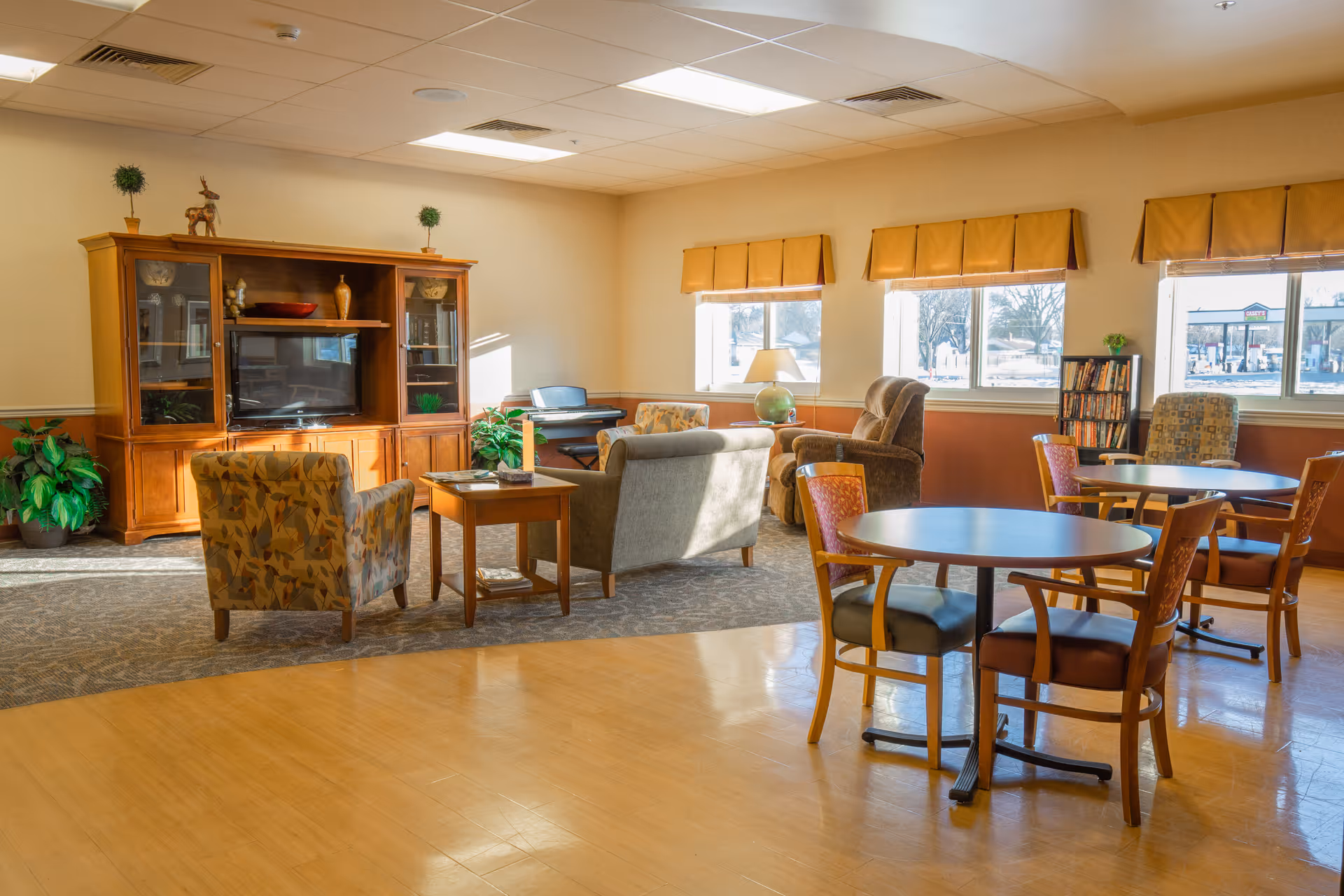 A bright and spacious common area in a senior living facility with several round tables and chairs on a wooden floor, a seating area with armchairs and a sofa on a carpet, a wooden entertainment center with a TV, and large windows with yellow valances letting in natural light.