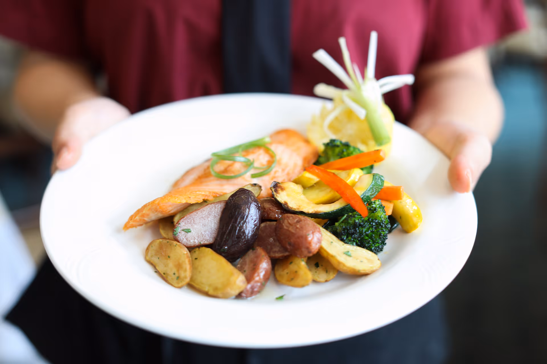 A person holding a white plate with a serving of cooked salmon, roasted potatoes, and mixed steamed vegetables including broccoli, zucchini, and red bell peppers, garnished with a lemon wedge and green onion curls.