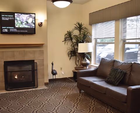 Cozy common room with a fireplace, wall-mounted TV, leather sofa, lamp, and large window.