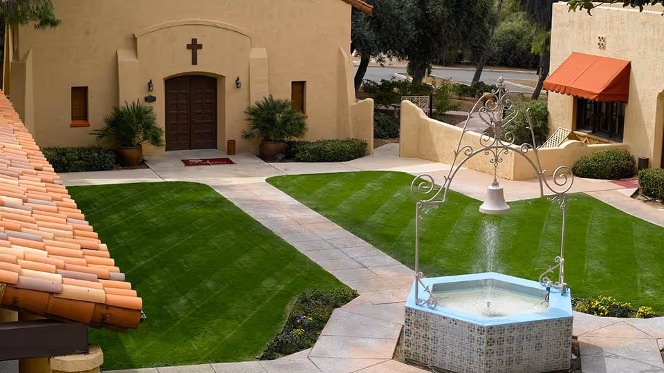 Courtyard with a central tiled fountain, striped green lawns and a stucco building entrance with a cross above the door.