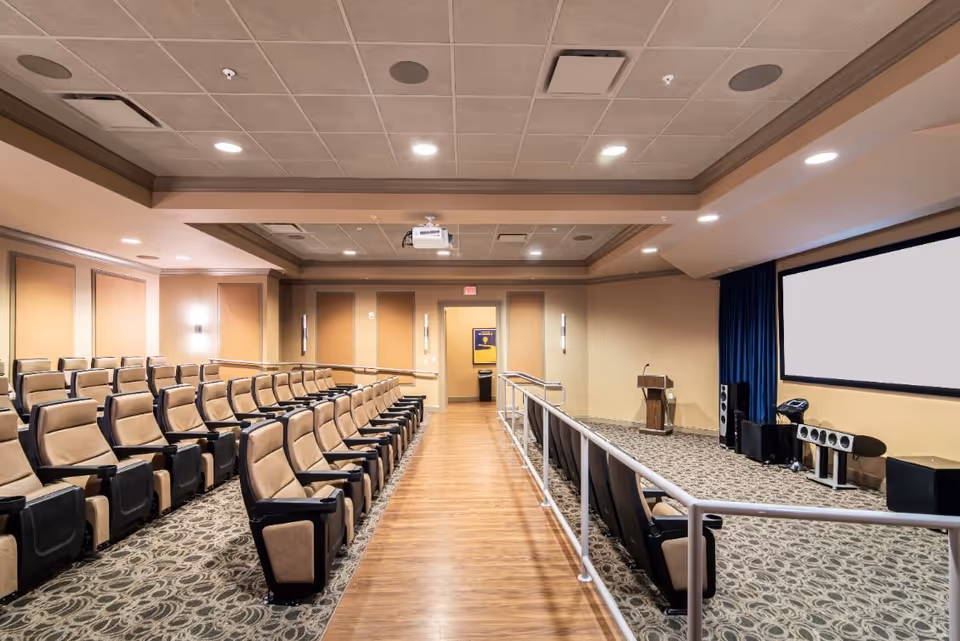 A spacious auditorium with rows of beige cushioned seats arranged in two sections separated by a central aisle with wooden flooring. The room has a patterned carpet, beige walls, a ceiling with recessed lighting, and a large projection screen at the front. There is a podium and audio equipment near the screen.