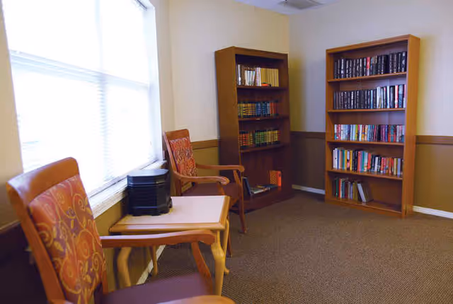 A small reading or sitting area with two wooden chairs featuring orange patterned cushions, a small wooden table between them, and two wooden bookshelves filled with books against the wall. The room has beige walls with a brown wainscot and a window with blinds letting in natural light.