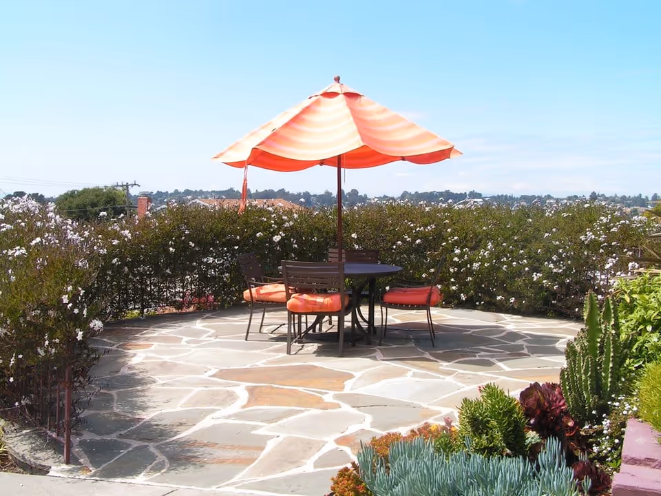 Stone patio with a round table, four chairs with orange cushions and a striped orange umbrella surrounded by flowering shrubs and succulents.