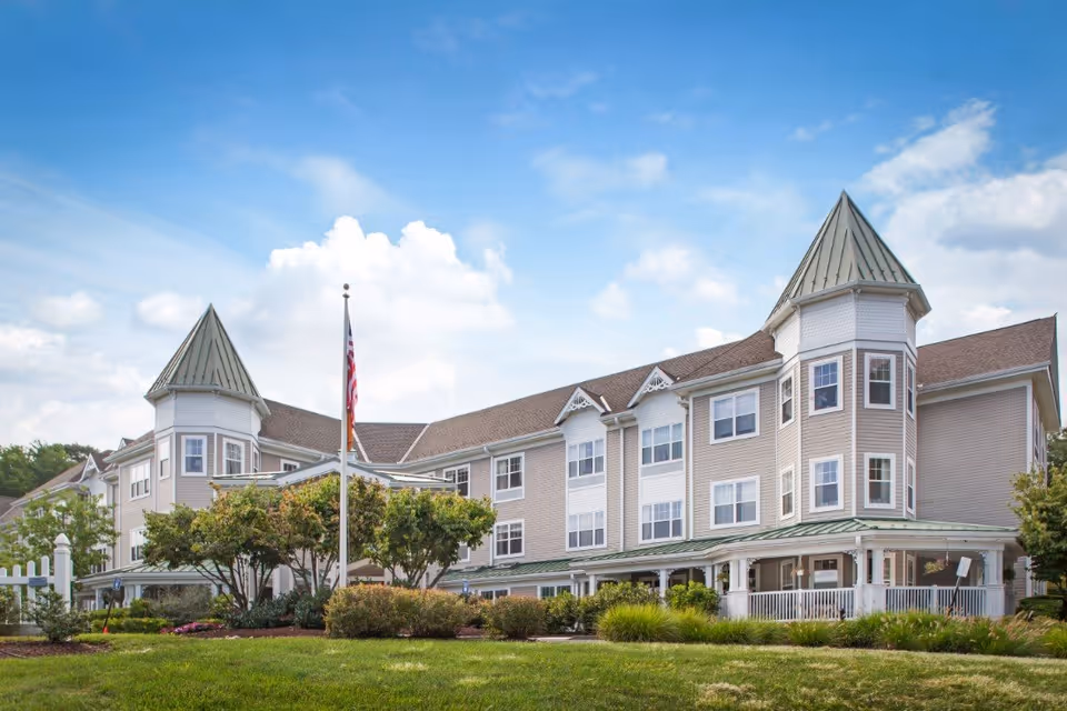 Exterior view of a large senior living facility building with beige siding, multiple windows, and two prominent turret-style roof sections. The building is surrounded by well-maintained landscaping, including bushes and trees, with an American flag on a flagpole in front. The sky is partly cloudy with blue patches.