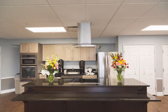 A modern communal kitchen with a large dark island, stainless steel appliances, range hood, and two vases of flowers on the counter.