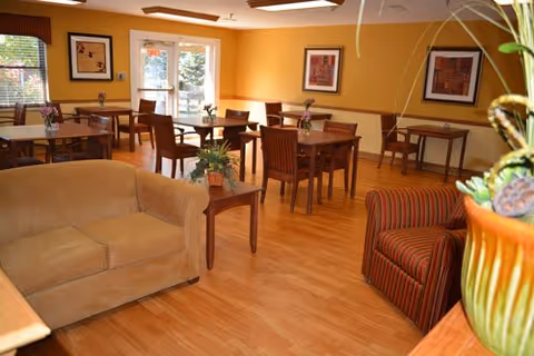 Bright communal dining room with wooden tables and chairs, upholstered sofas and armchairs, framed artwork, and potted plants.