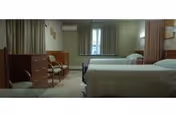 A senior living community bedroom with two single beds, each with white bedding. There are chairs and a wooden dresser on the left side of the room, and a window with curtains in the center background. The room has soft lighting and neutral-colored walls.