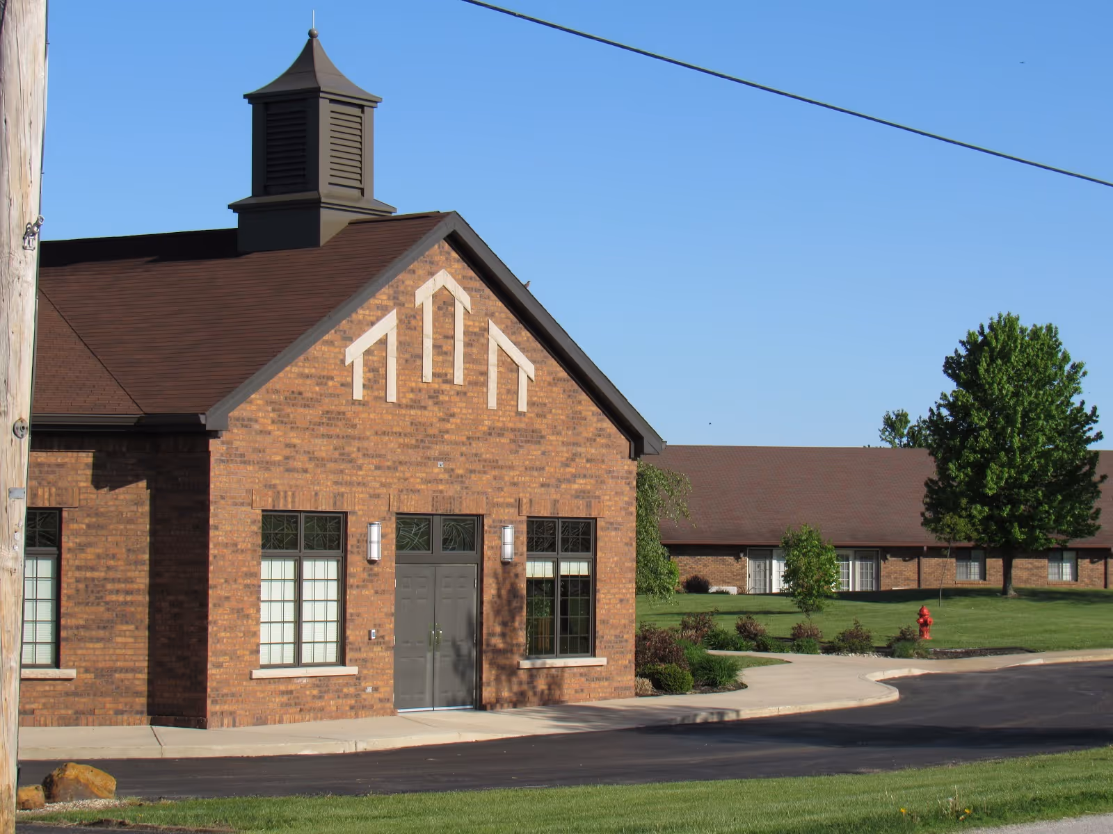 Brick-front entrance of a single-story building with a cupola, windows, driveway, landscaped lawn and clear blue sky.