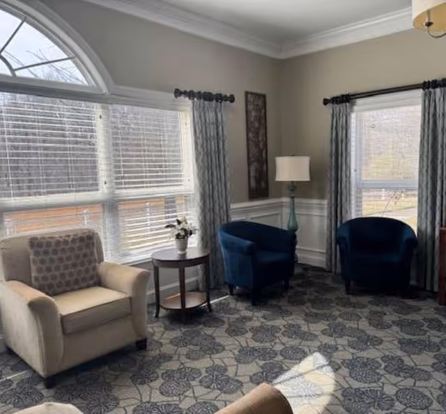 A cozy sitting area with three armchairs, one beige and two dark blue, arranged near large windows with white blinds and patterned curtains. A small round wooden table with a flower vase is placed between the beige and one blue chair. A floor lamp with a white shade stands in the corner, and the room has a patterned carpet and light-colored walls with white trim.