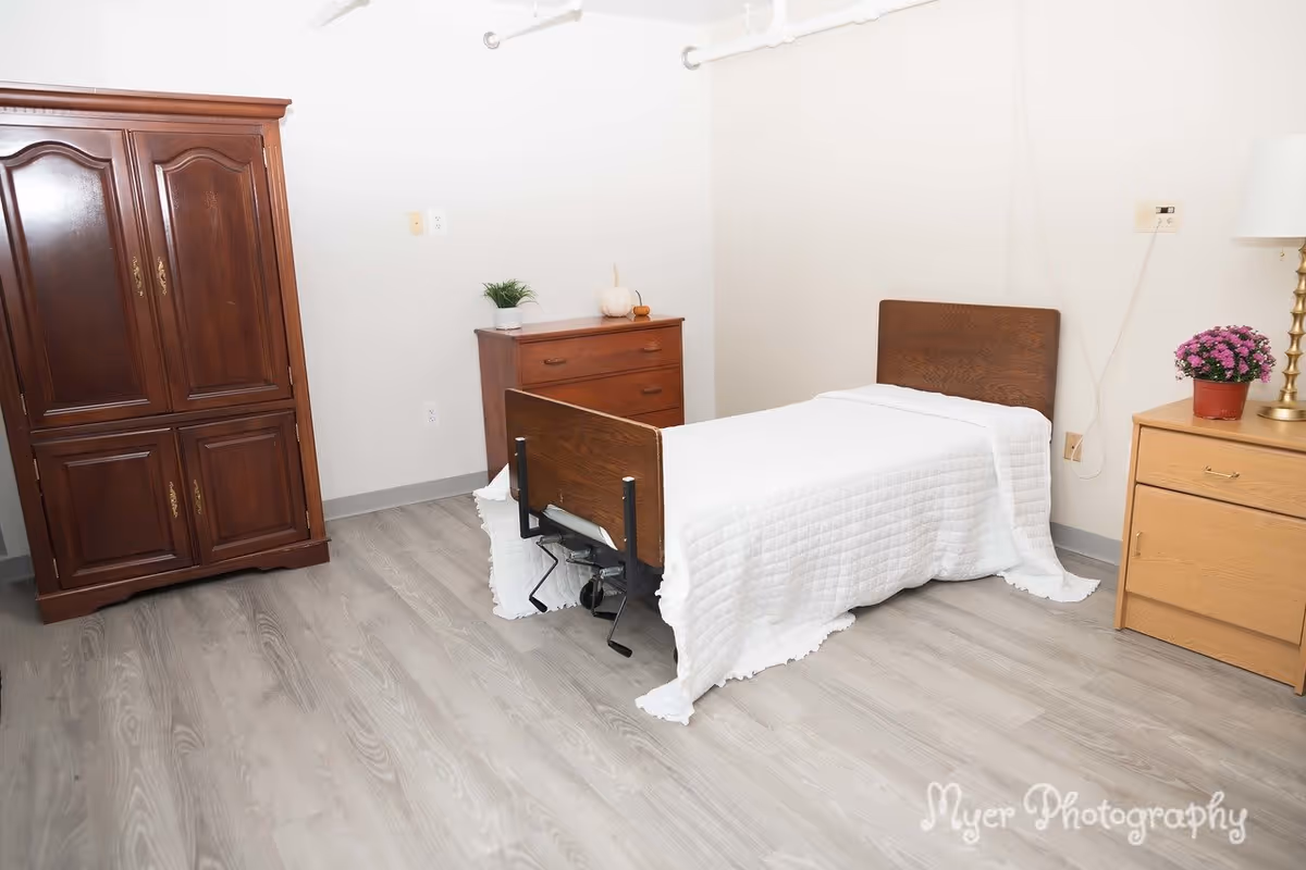 A simple bedroom with a single bed covered in a white quilt, a wooden wardrobe, a wooden dresser with a small plant and decorative items, and a wooden nightstand with a lamp and a potted flower. The room has light-colored walls and wood-patterned flooring.
