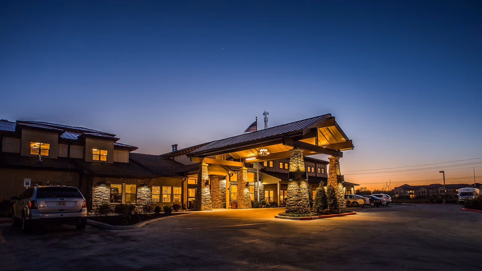 Exterior view of Vitality Court senior living facility at dusk, showing the main entrance with stone pillars and warm lighting, cars parked in the parking lot, and a clear sky transitioning from sunset to night.