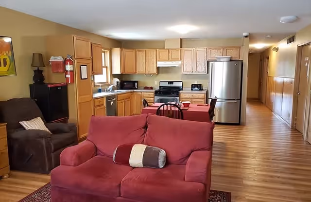 Interior view of a senior living facility showing a combined living room and kitchen area. The living room has a red sofa with a decorative pillow and a brown armchair with a cushion. The kitchen features wooden cabinets, a stainless steel refrigerator, a stove, a microwave, and a dishwasher. A small dining table with chairs is placed between the living room and kitchen. The floor is wooden, and the walls are painted light beige. There is a hallway visible to the right.