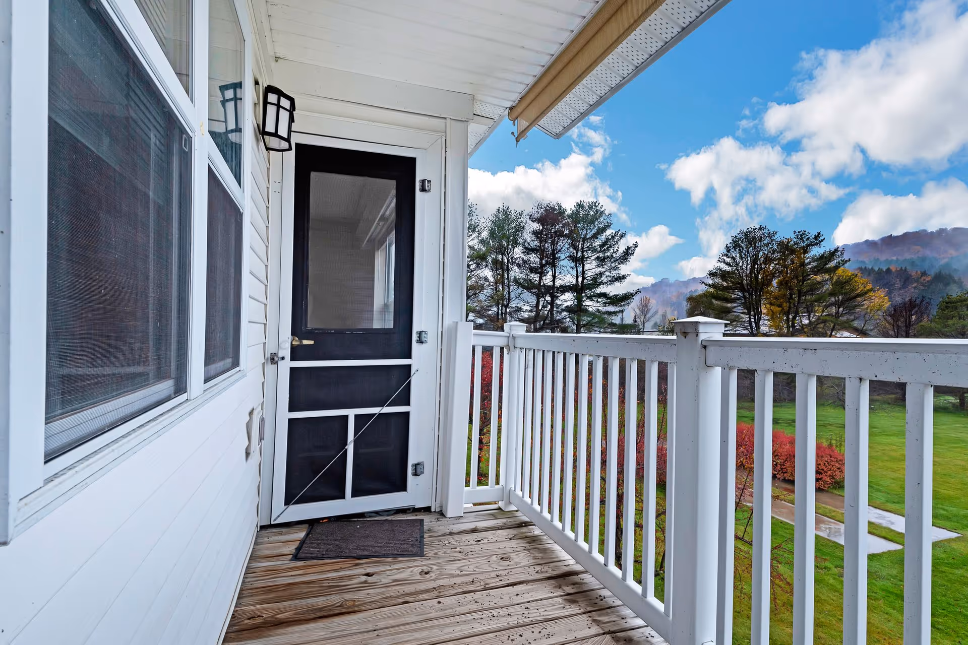 View of a small wooden balcony with white railing and a black screen door leading inside. The balcony overlooks a green lawn with trees and bushes under a partly cloudy blue sky.