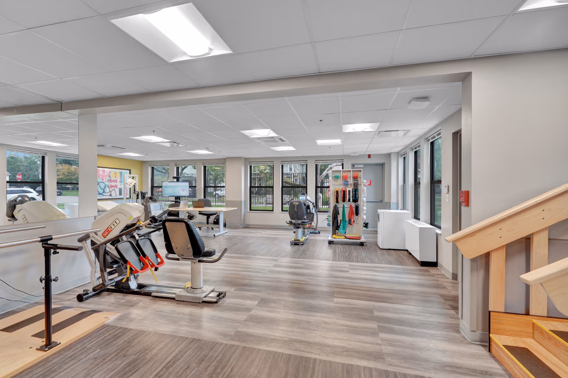 A spacious rehabilitation and exercise room with large windows letting in natural light. The room features exercise equipment including a seated leg press machine and a recumbent bike, parallel bars for walking practice, a rack with colorful resistance bands, and a wooden staircase for physical therapy. The flooring is a mix of wood and vinyl, and the ceiling has recessed lighting.