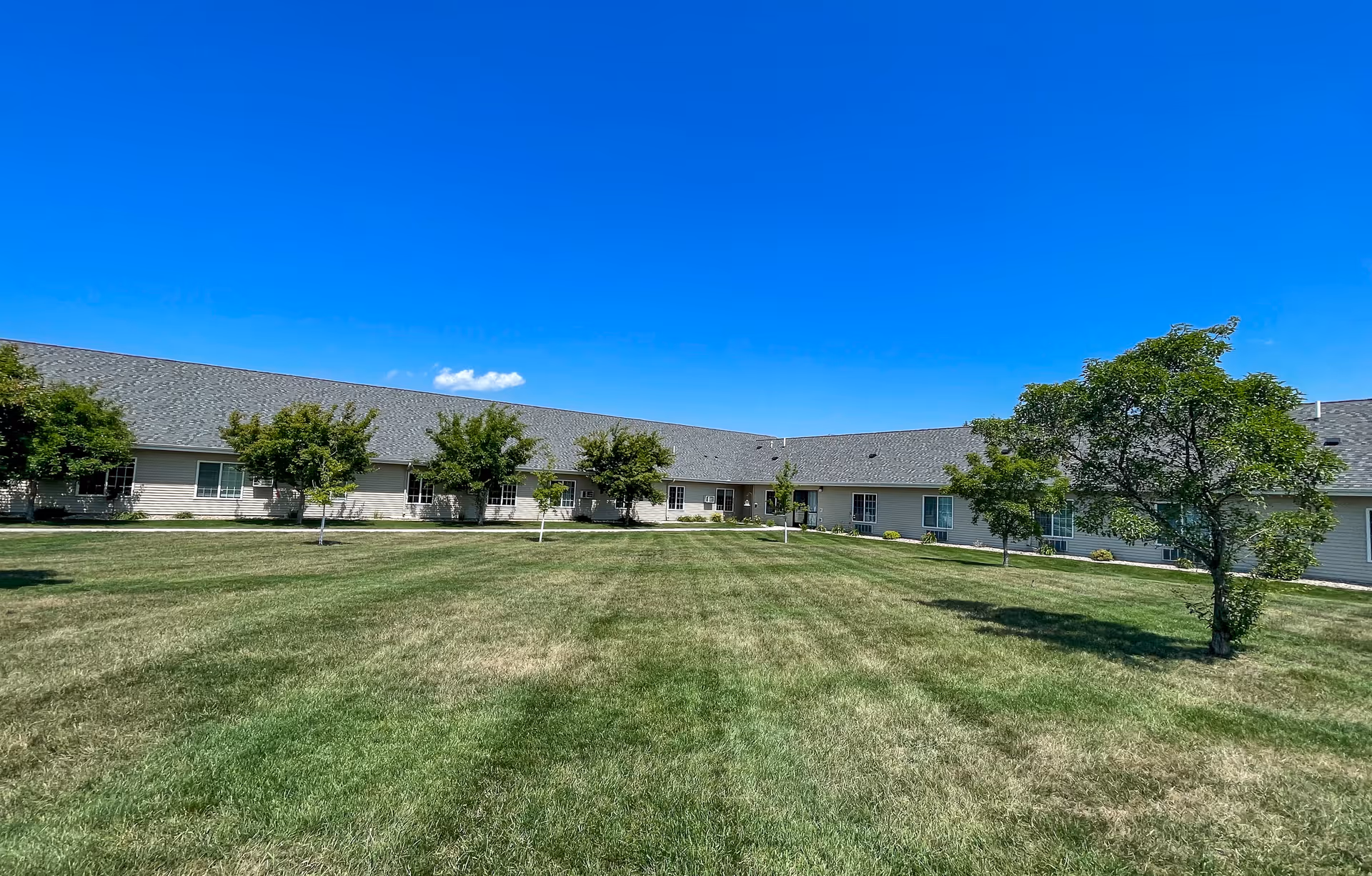 Wide grassy courtyard with small trees in front of a long single-story residential building under a clear blue sky.