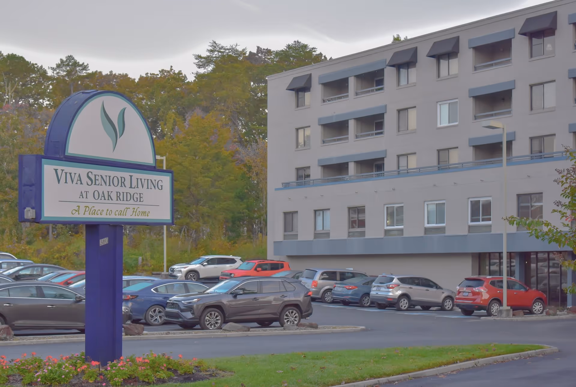 Exterior view of a senior living facility named Viva Senior Living at Oak Ridge, showing a multi-story building with balconies and a parking lot filled with cars. There is a large sign in the foreground with the facility's name and the tagline 'A Place to call Home'. Trees and greenery are visible in the background.