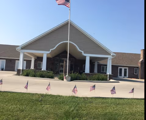 Front exterior view of a single-story building with a large covered entrance supported by white columns. An American flag is flying on a tall flagpole in front of the entrance, and several smaller American flags are lined up along the edge of the grass near the driveway.