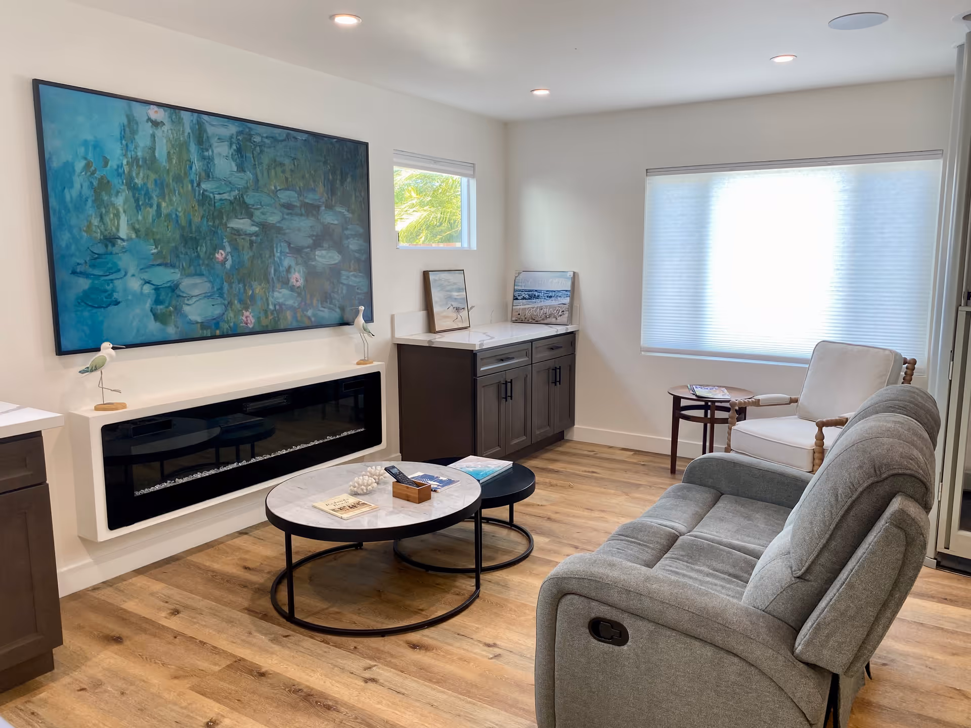 Bright modern living room with a gray reclining sofa, armchair, round coffee tables, a wall-mounted electric fireplace, and a large blue-green painting.