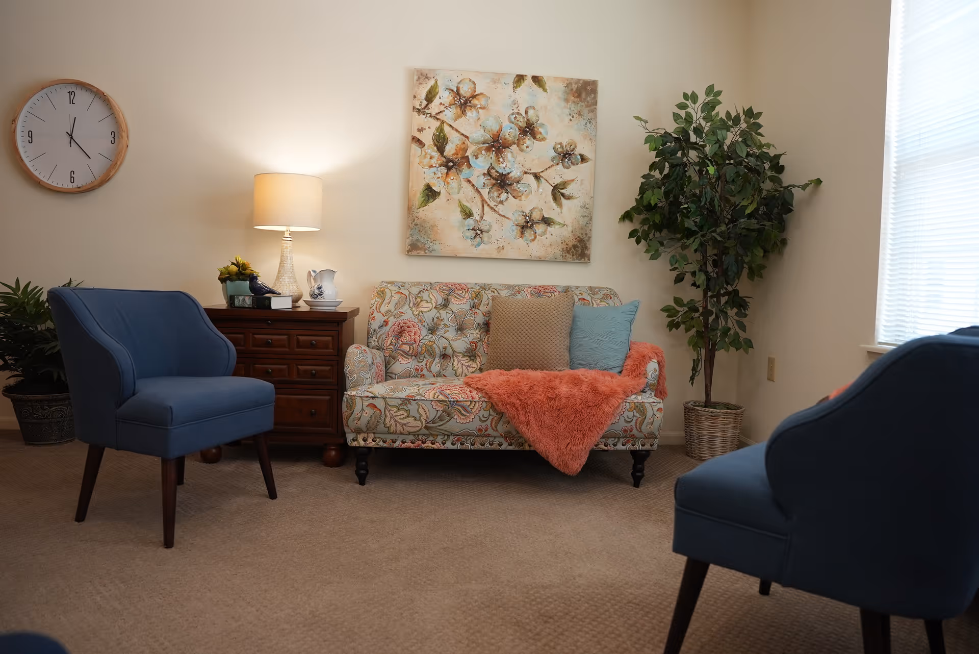 A cozy living room with a floral patterned loveseat adorned with three pillows and a coral throw blanket. Two blue upholstered chairs face the loveseat. A wooden side table with a lamp, a small plant, and a decorative pitcher sits next to the loveseat. A large leafy plant is in the corner near a window with blinds. A round wall clock and a floral painting hang on the beige walls.