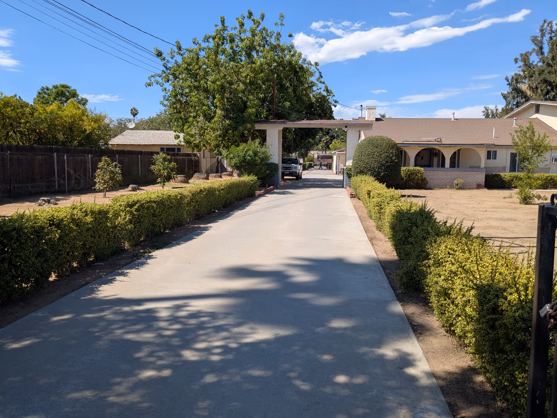 Driveway lined with hedges leading to a single-story building with an arched porch under a blue sky.