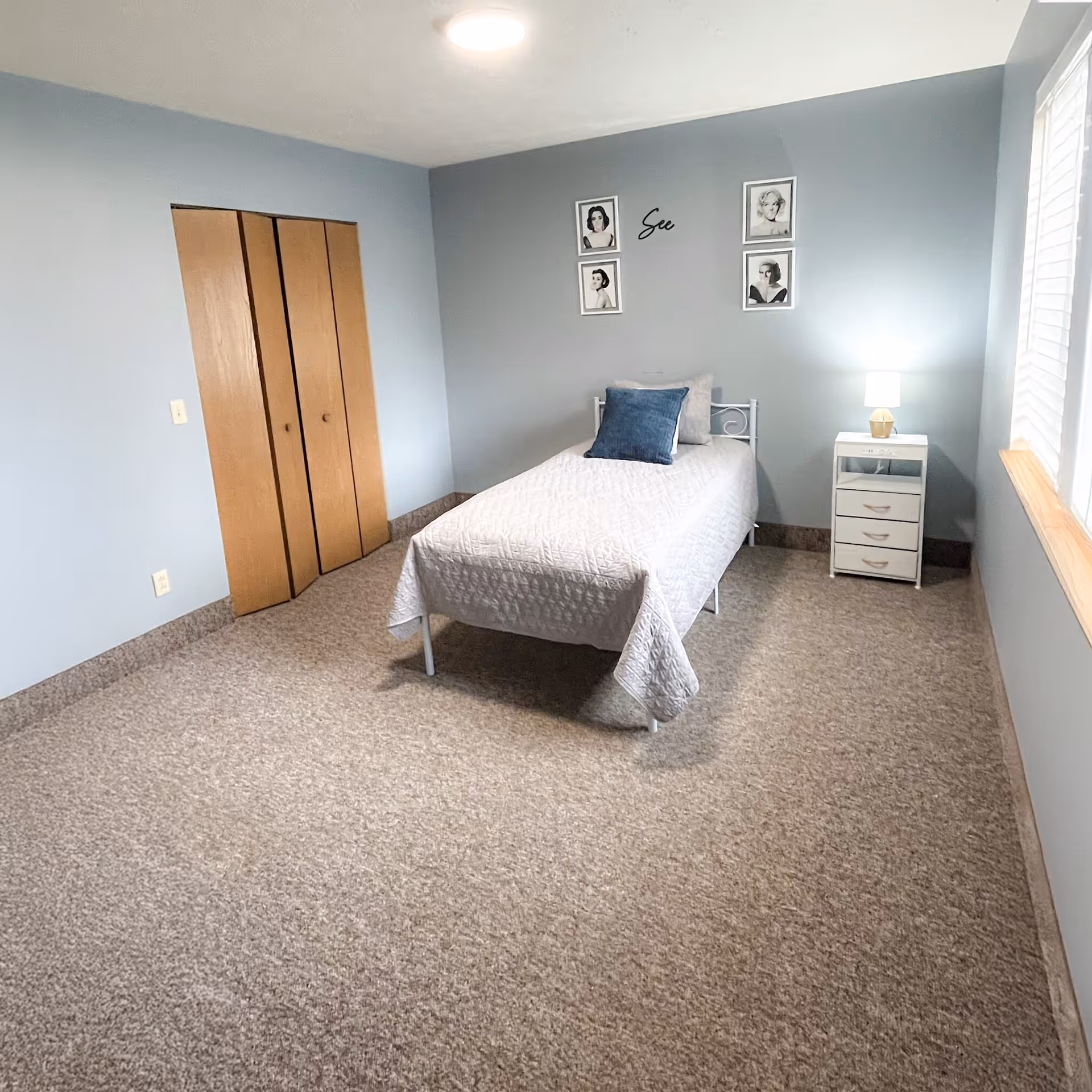 Simple single bedroom with a twin bed, nightstand and lamp, closet doors, framed wall art, and carpeted floor.