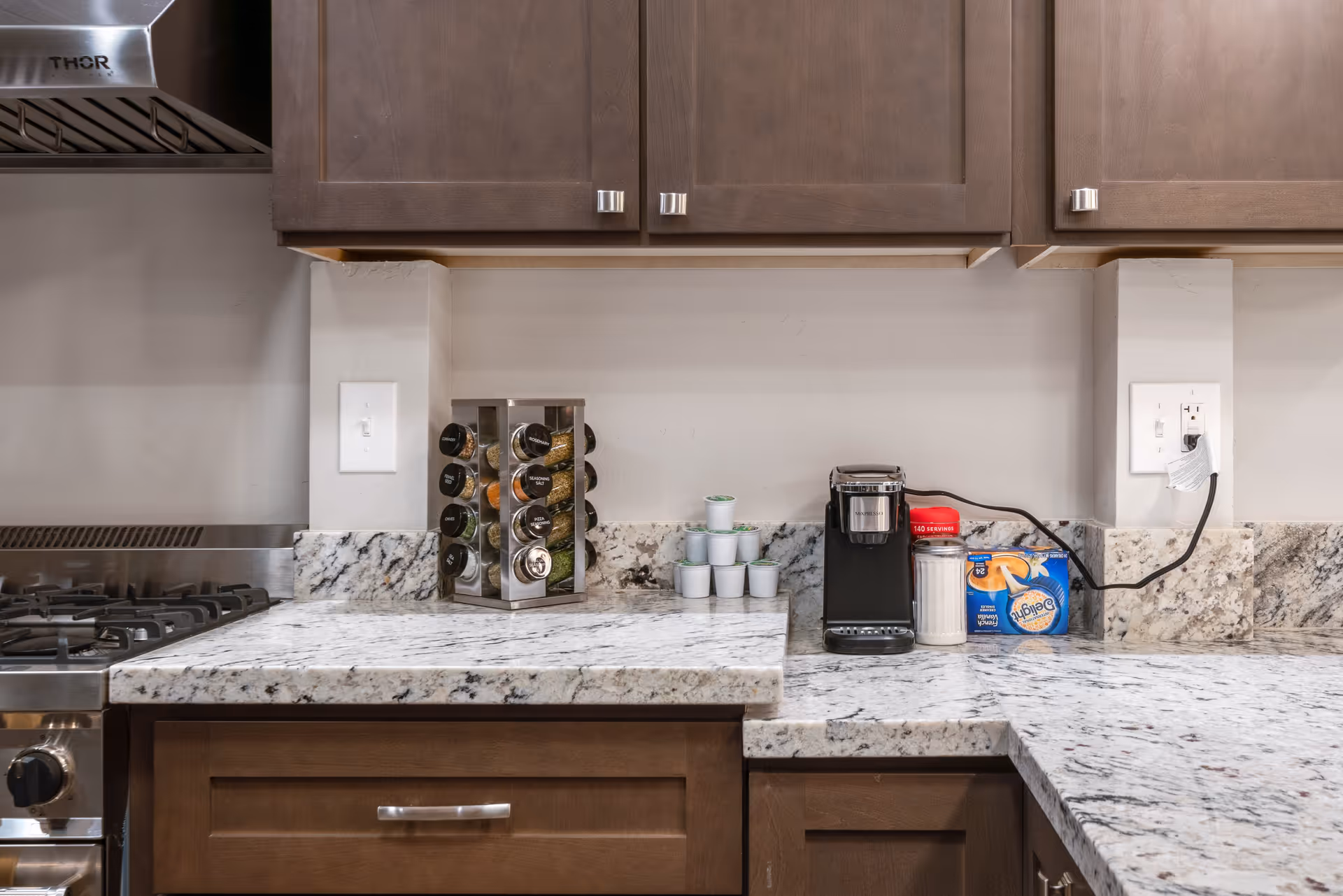 A kitchen countertop with a stainless steel stove on the left, a rotating spice rack with various spices, a stack of coffee pods, a black single-serve coffee maker, a container of sugar, and a box of Delights French Vanilla coffee pods. The countertop is granite with brown wooden cabinets above and below.