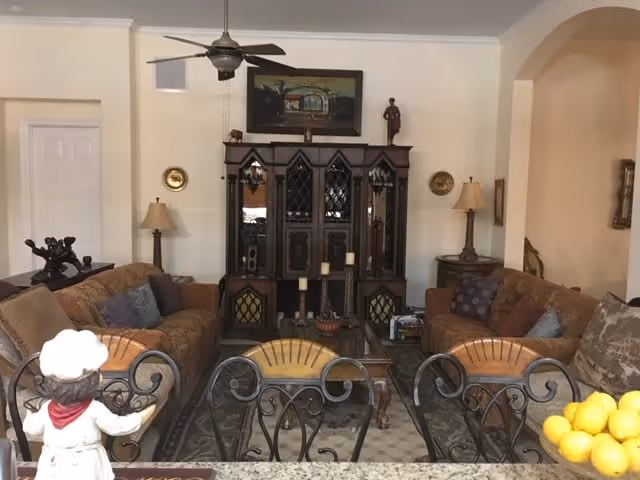 Living room with two sofas facing an ornate wooden cabinet, a ceiling fan, lamps, decorative chairs in the foreground, and a coffee table.