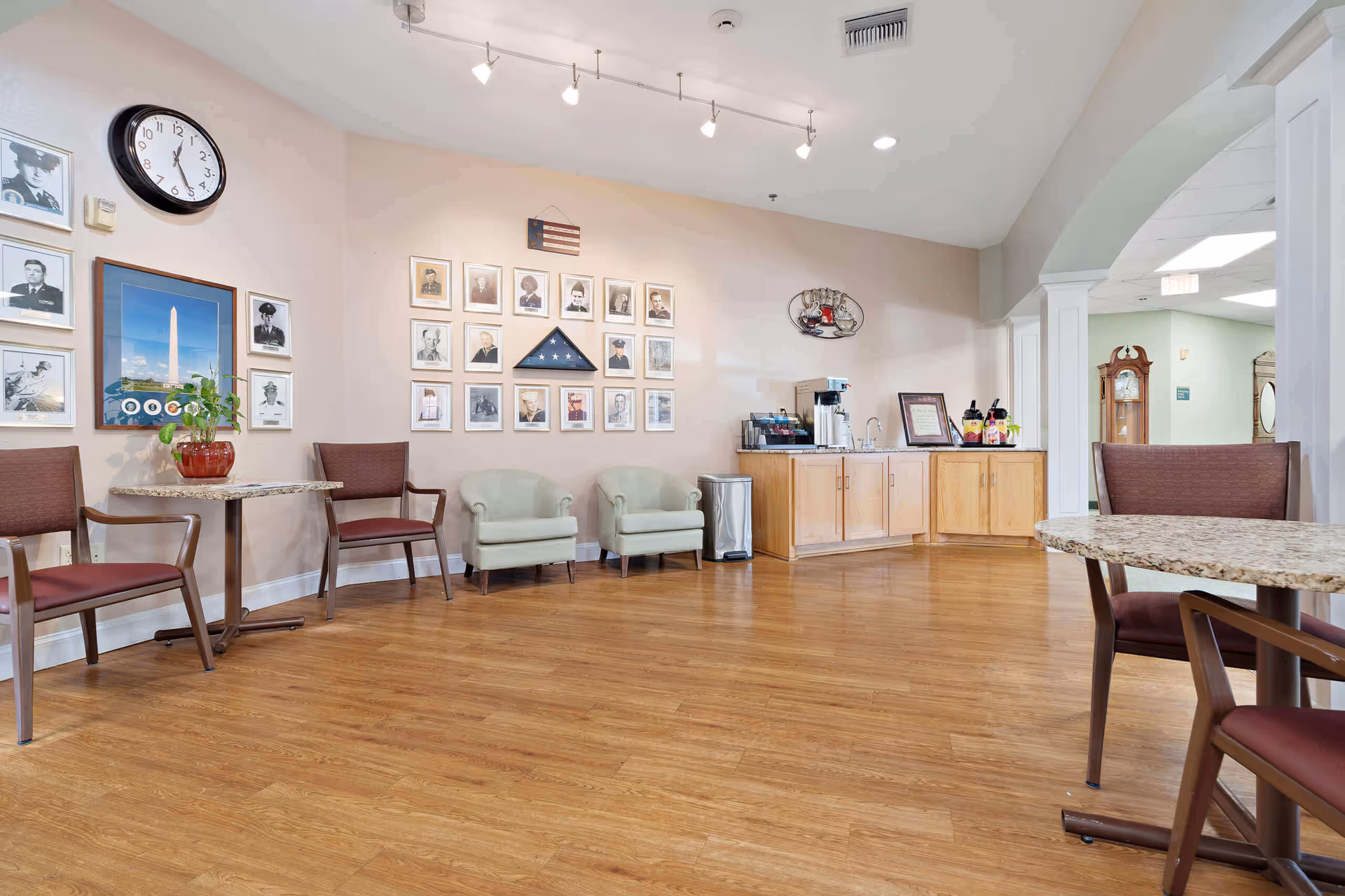 A seating area in a senior living facility with wooden flooring, several chairs, a small table with a plant, and a wall decorated with framed photographs and a folded American flag. There is a coffee station with a coffee maker and supplies on a wooden cabinet. The space is well-lit with ceiling lights and has an archway leading to another room.