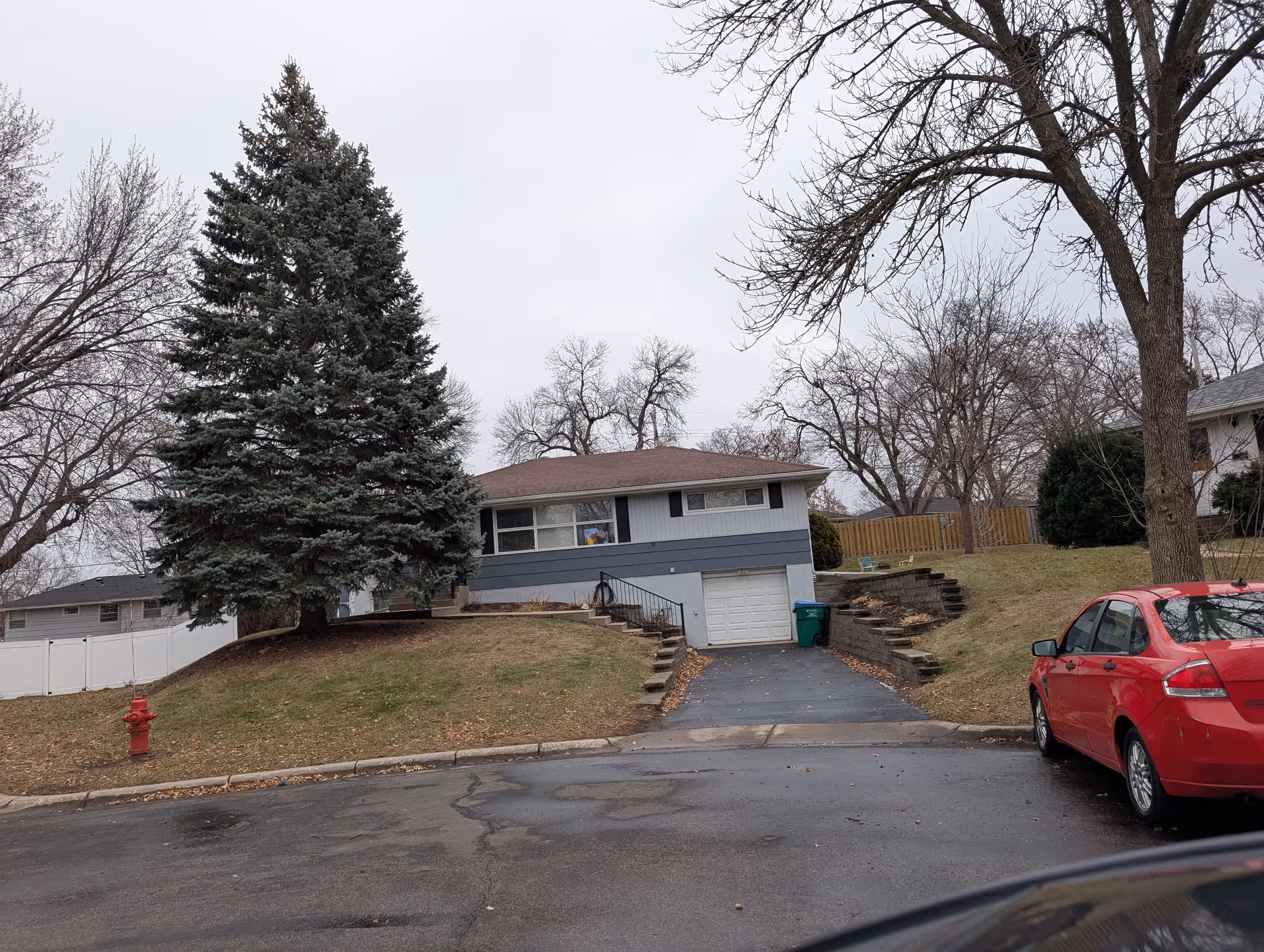 A single-story residential house with a sloped driveway leading to a garage. The house has a brown roof and blue-gray siding with white trim. There is a large evergreen tree on the left side of the front yard and a red car parked on the street to the right. Leafless deciduous trees surround the property, and a red fire hydrant is visible near the curb on the left.