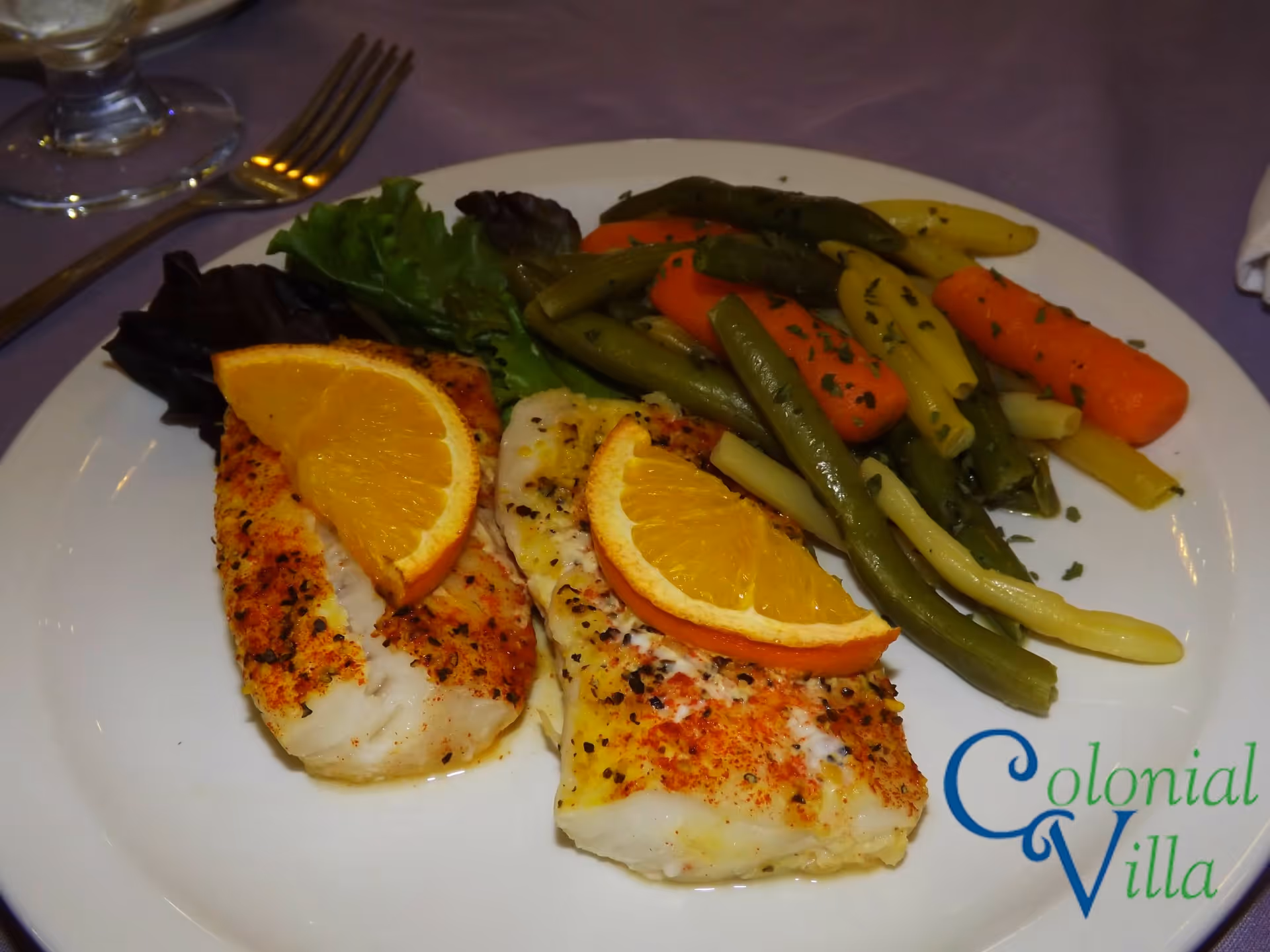 A plate of seasoned cooked fish fillets topped with orange slices, served with steamed mixed vegetables including green beans, yellow beans, and carrots, accompanied by leafy greens on the side. A fork and a glass of water are partially visible on a purple tablecloth.