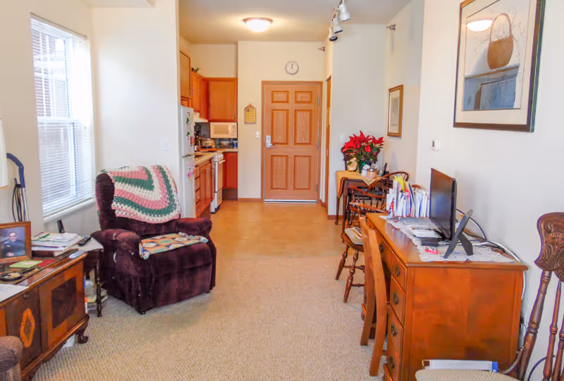 Cozy living area with a recliner, wooden furniture and desk, a small kitchen to the left, and the entrance door at the far end.