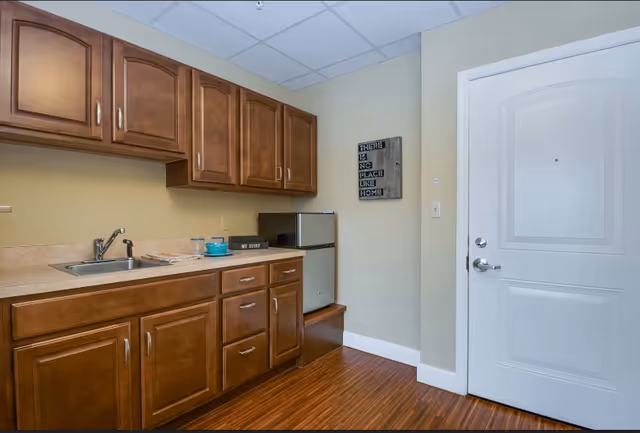 Interior view of a small kitchenette area with wooden cabinets, a countertop with a sink, a mini refrigerator, and a white door. There is a decorative sign on the wall that reads 'There is no place like home.'