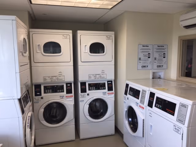 Laundry room with multiple stacked and side-by-side commercial washing machines and dryers. The machines are white and have digital control panels. There are service instruction signs on the wall.