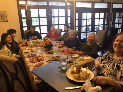 A group of elderly people and a few younger adults sitting around a dining table enjoying a meal together in a room with large windows and wooden framed glass doors in the background.