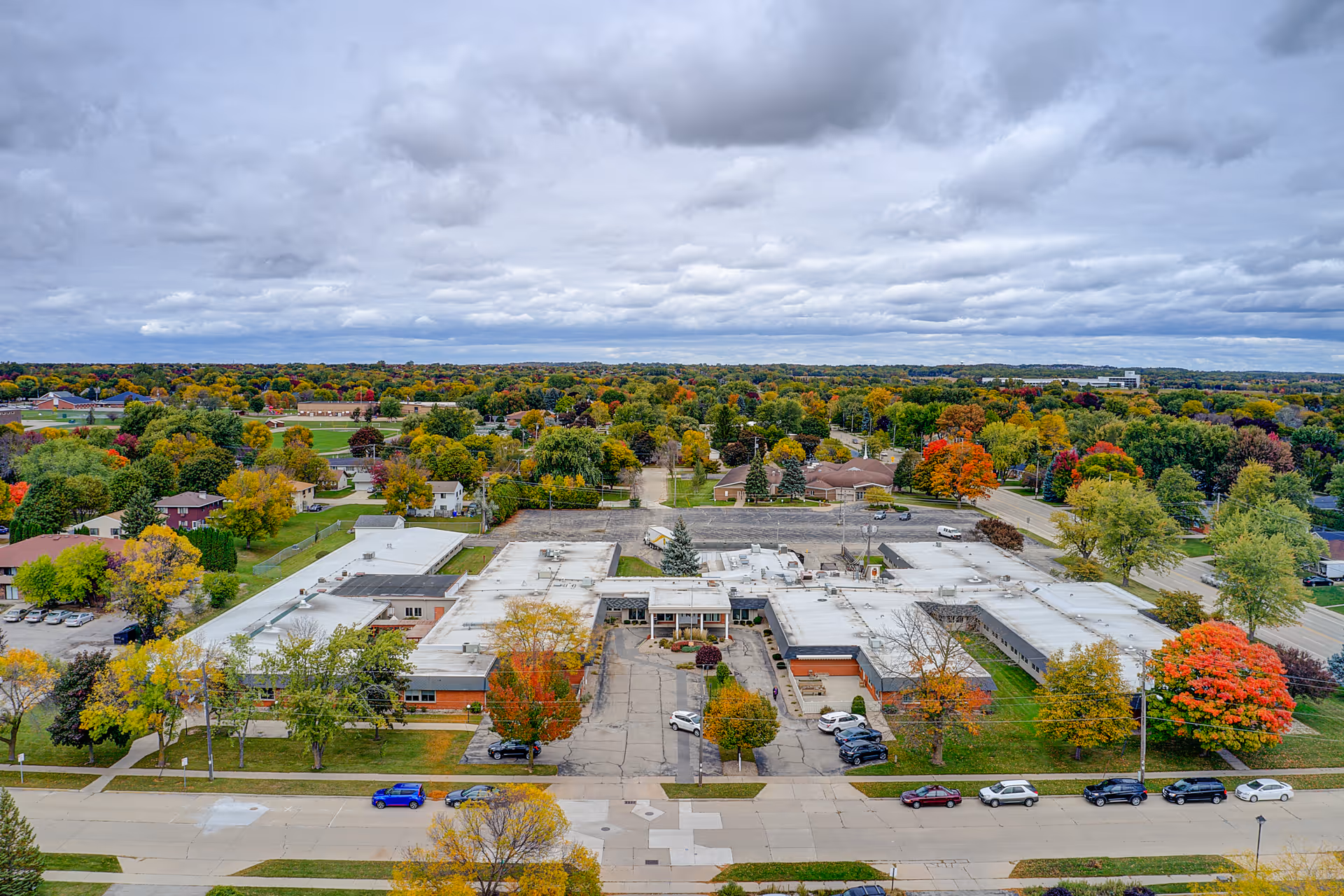 Aerial view of a large single-story building with a white roof surrounded by trees with autumn foliage. Several cars are parked in front of the building along the street. The sky is cloudy and the area appears to be a suburban neighborhood with many houses and green spaces.