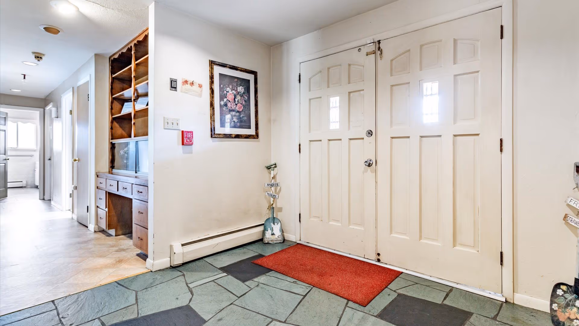 Interior view of an entryway with double cream-colored doors, a red doormat, and a stone tile floor. To the left, there is a built-in wooden shelving unit with drawers and cubbies, and a hallway leading to other rooms. A framed floral picture hangs on the wall above a fire alarm.