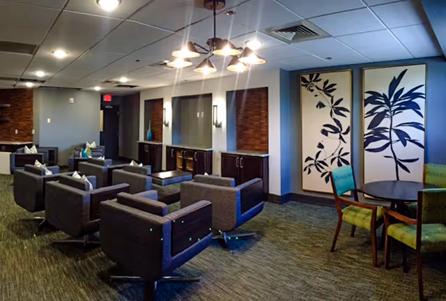A modern lounge area with multiple gray armchairs arranged in rows facing a wall with mirrors and cabinets. To the right, there is a round table with green chairs and two large decorative panels featuring black leaf designs on a white background. The ceiling has recessed lighting and a central chandelier with multiple bulbs.