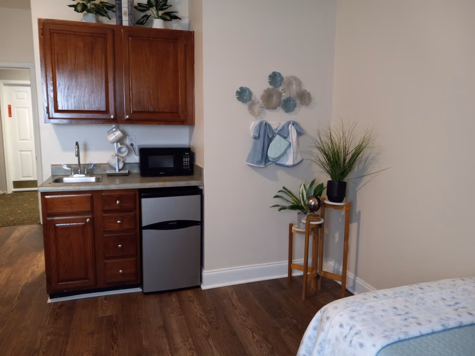 Small in-room kitchenette with wooden cabinets, sink, microwave, mini-fridge and decorative plants beside a bed.