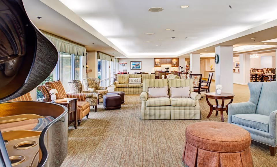 A spacious and well-lit living room area in a senior living facility featuring a grand piano on the left, multiple comfortable armchairs and sofas arranged around small tables, large windows with curtains allowing natural light, and a dining area visible in the background.