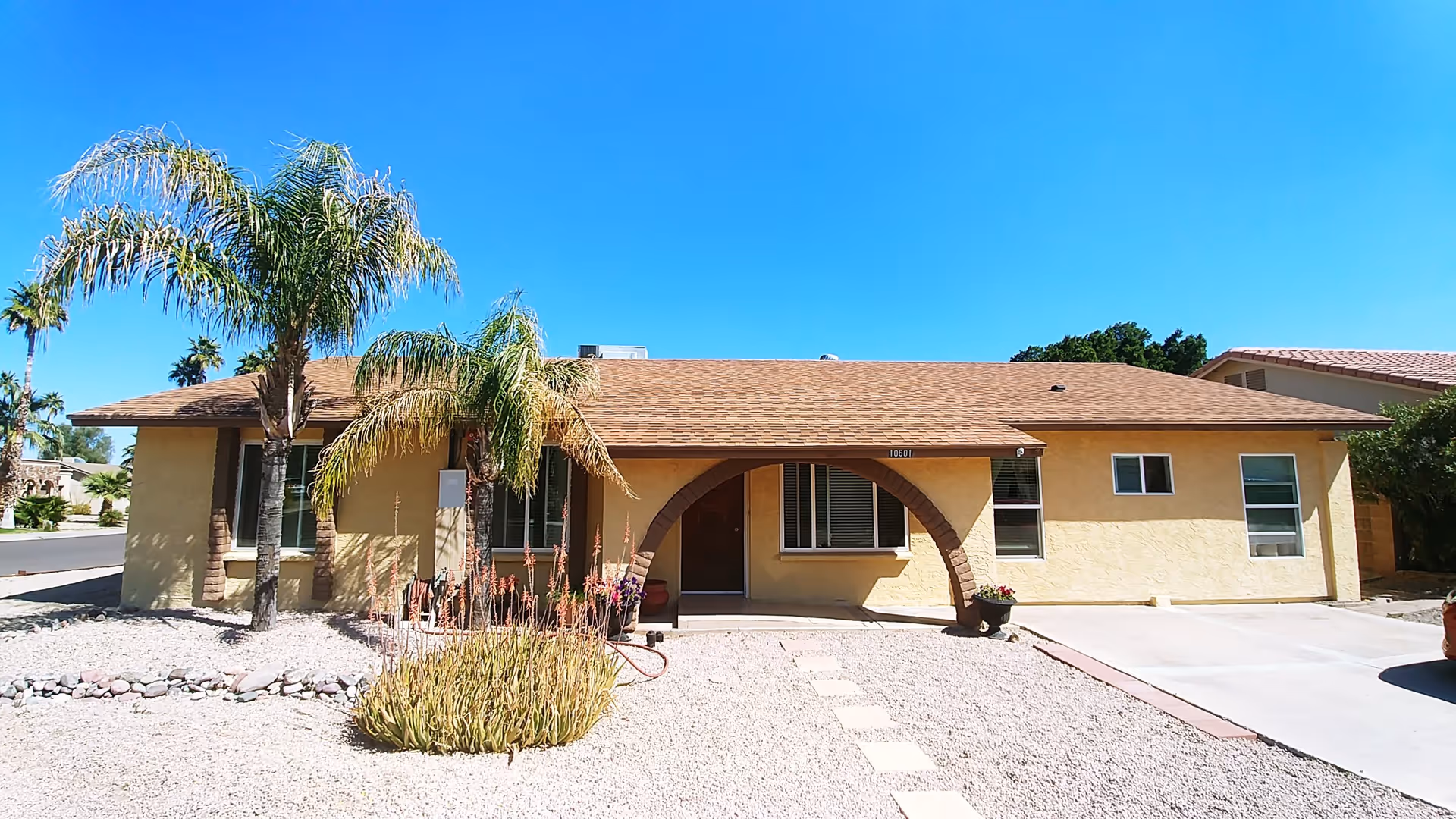 Single-story yellow stucco house with a brown shingle roof, an arched entrance, and several windows. The front yard is landscaped with gravel, a few palm trees, and desert plants. The sky is clear and blue.