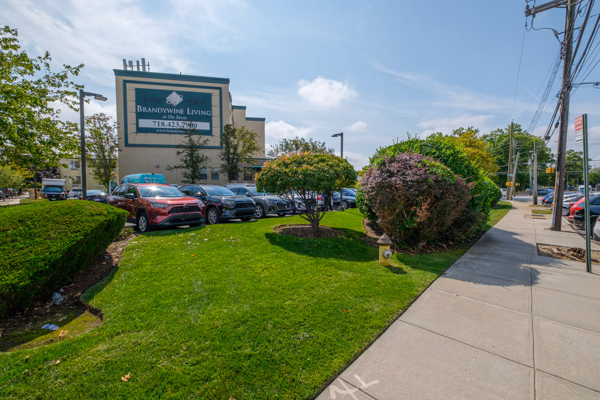 Exterior view of Brandywine Living at The Savoy building with a parking lot filled with cars, green grass, trimmed bushes, a sidewalk, and a clear sky.