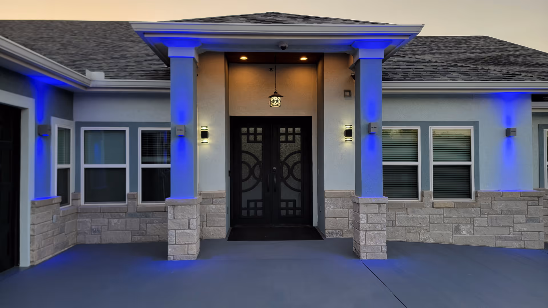Front entrance of a building with double black doors featuring decorative glass panels, flanked by two columns with stone bases. The exterior walls are light blue with stone accents, and blue lights illuminate the columns and walls. There are four windows with white frames and blinds on either side of the entrance. A hanging lantern light is above the door, and two wall-mounted lights are on either side of the door.