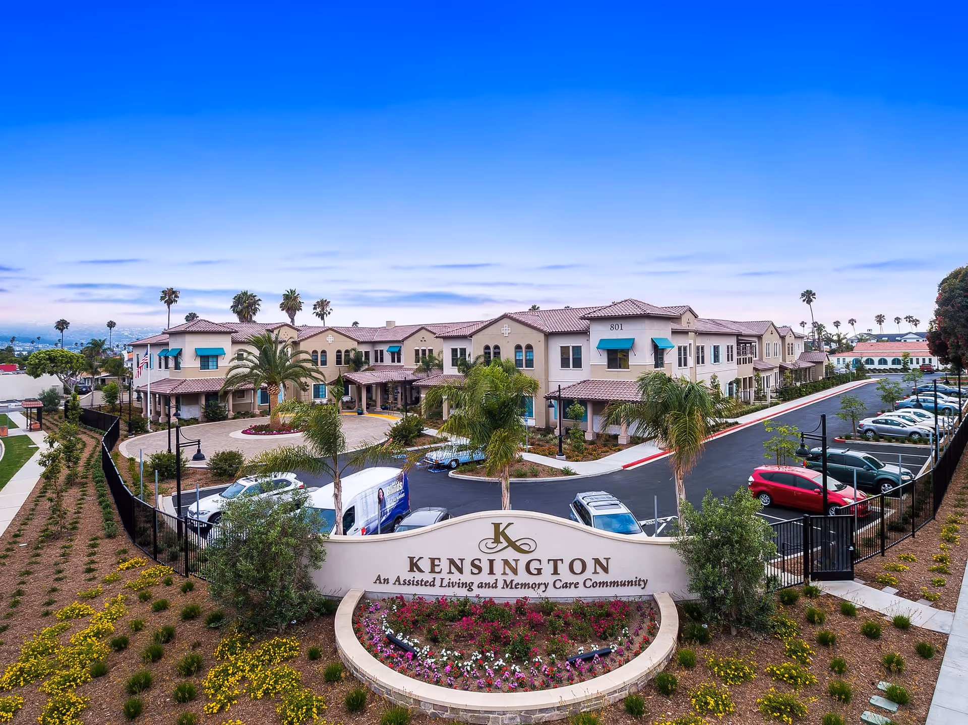 Exterior view of The Kensington Redondo Beach, an assisted living and memory care community, showing a large two-story building with beige walls and teal awnings. The foreground features a landscaped garden with flowers and palm trees, a curved driveway with parked cars, and a prominent sign displaying the community's name.