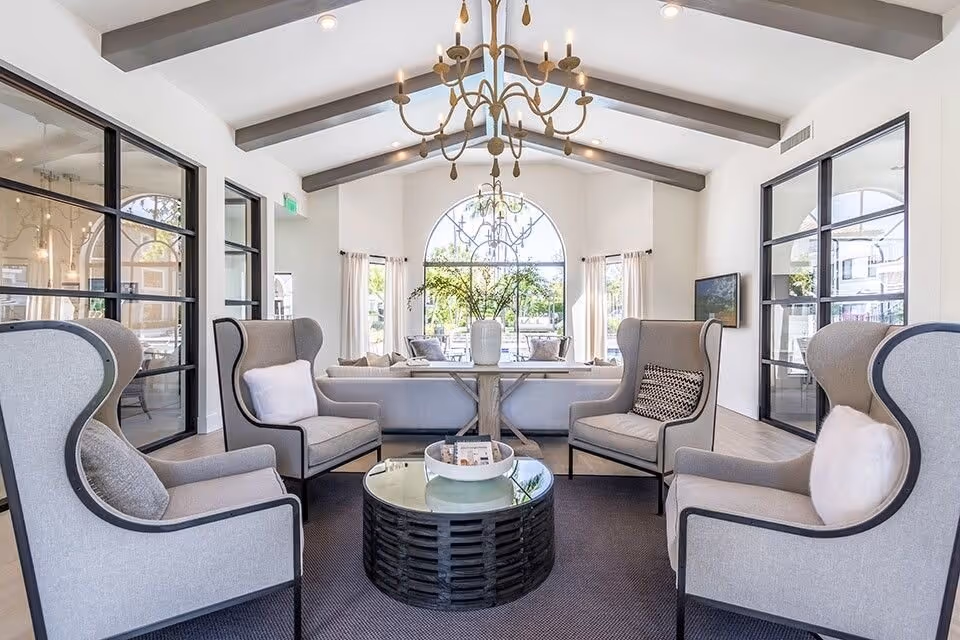 Bright, airy lounge with a high beamed ceiling and chandelier, four wingback chairs around a round coffee table facing a sofa and large arched window.