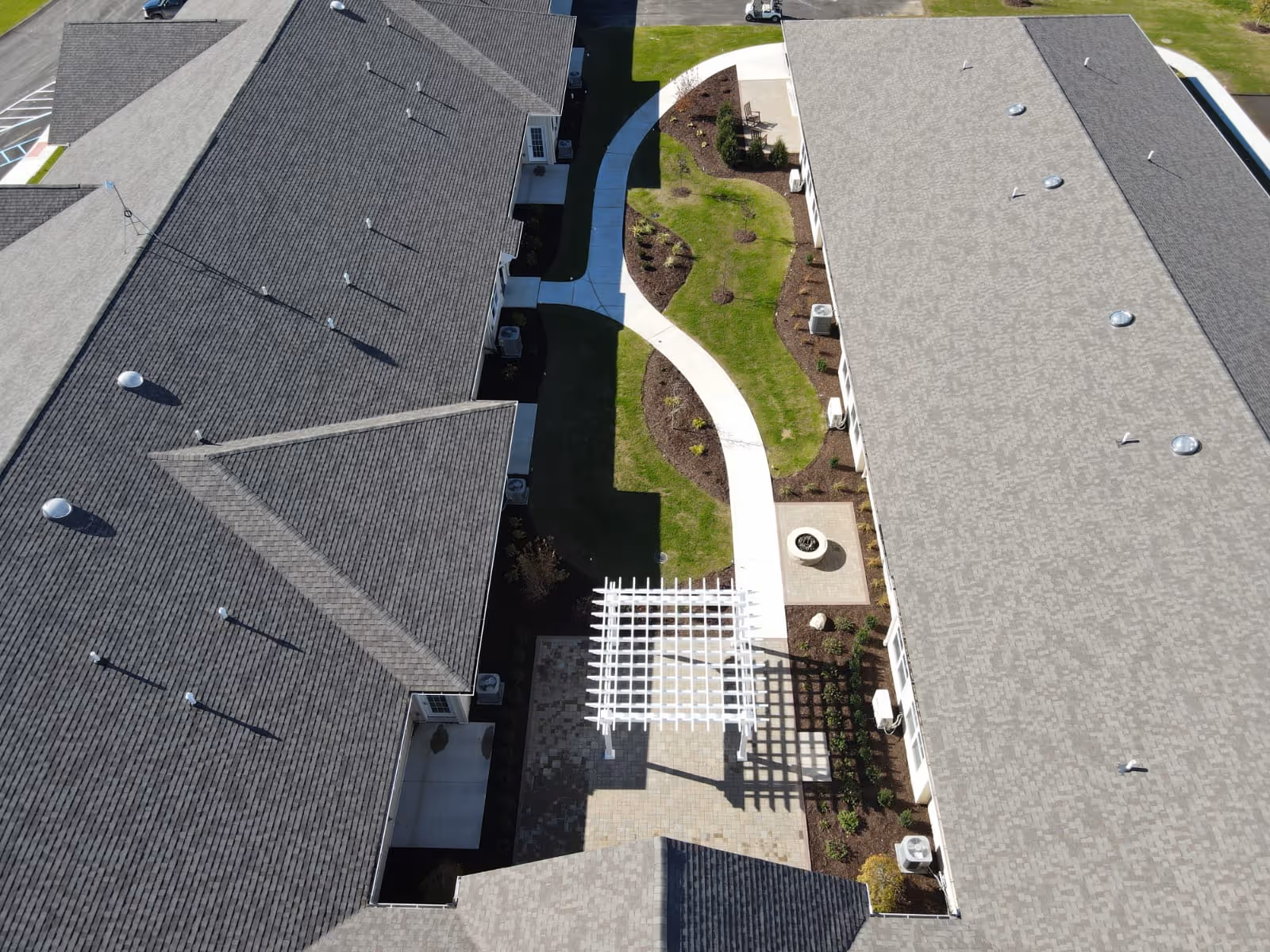 Aerial view of a landscaped courtyard with a winding sidewalk and pergola between two long building roofs.