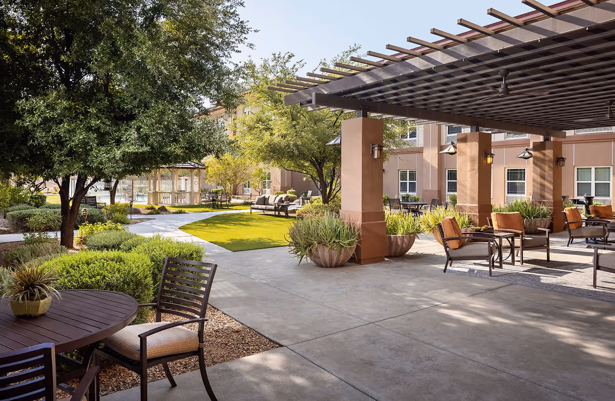Outdoor patio area at Belmont Village Senior Living Scottsdale featuring seating arrangements with chairs and tables under a pergola, surrounded by trees, shrubs, and a well-maintained garden with a gazebo in the background.