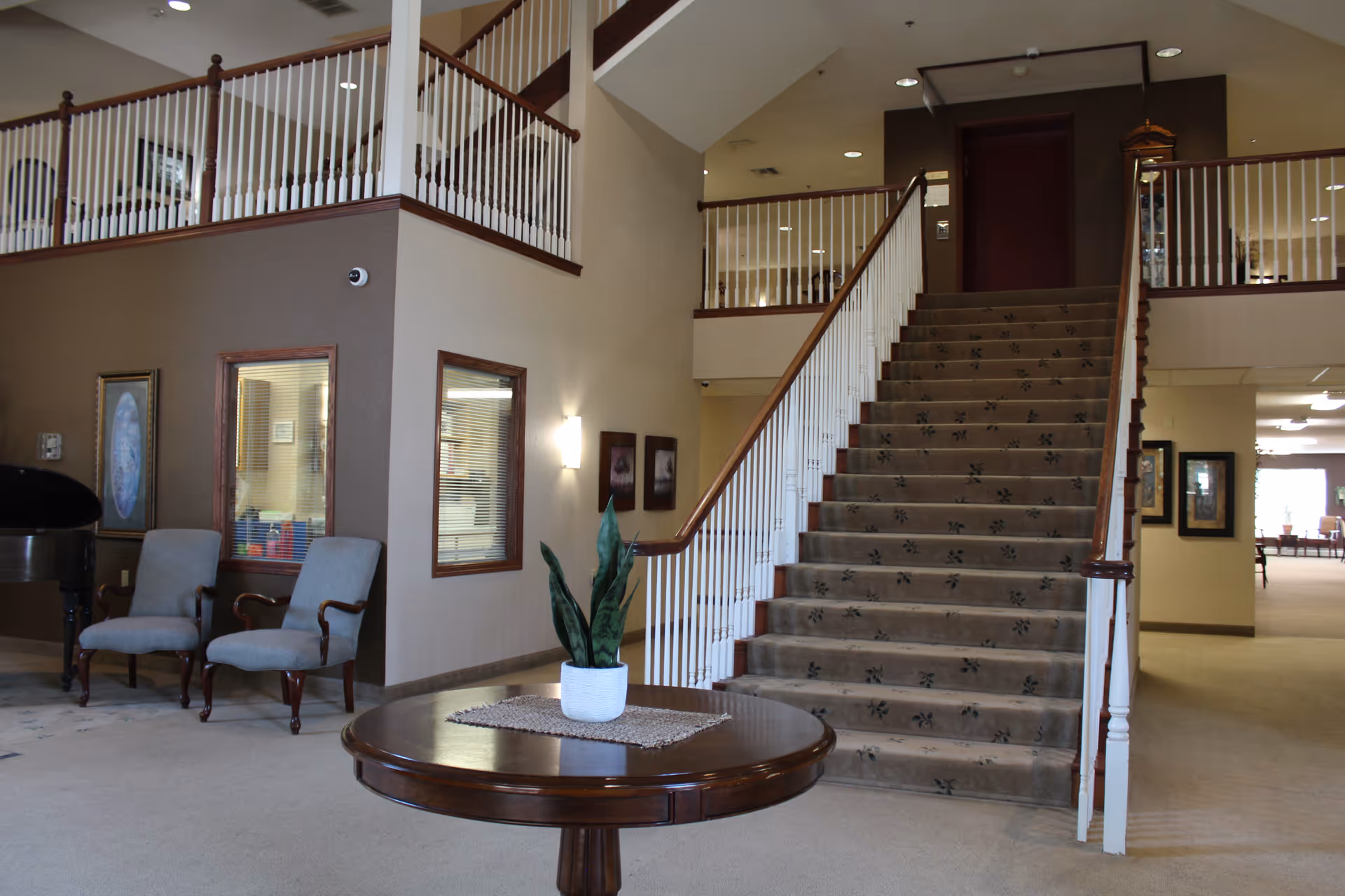 Interior view of a senior living facility showing a carpeted staircase with white railings and wooden handrails. In front of the staircase is a round wooden table with a potted plant on a woven mat. To the left, there are two upholstered chairs and a piano. The walls are beige with framed artwork and windows with blinds. The area is well-lit with ceiling lights and wall sconces.