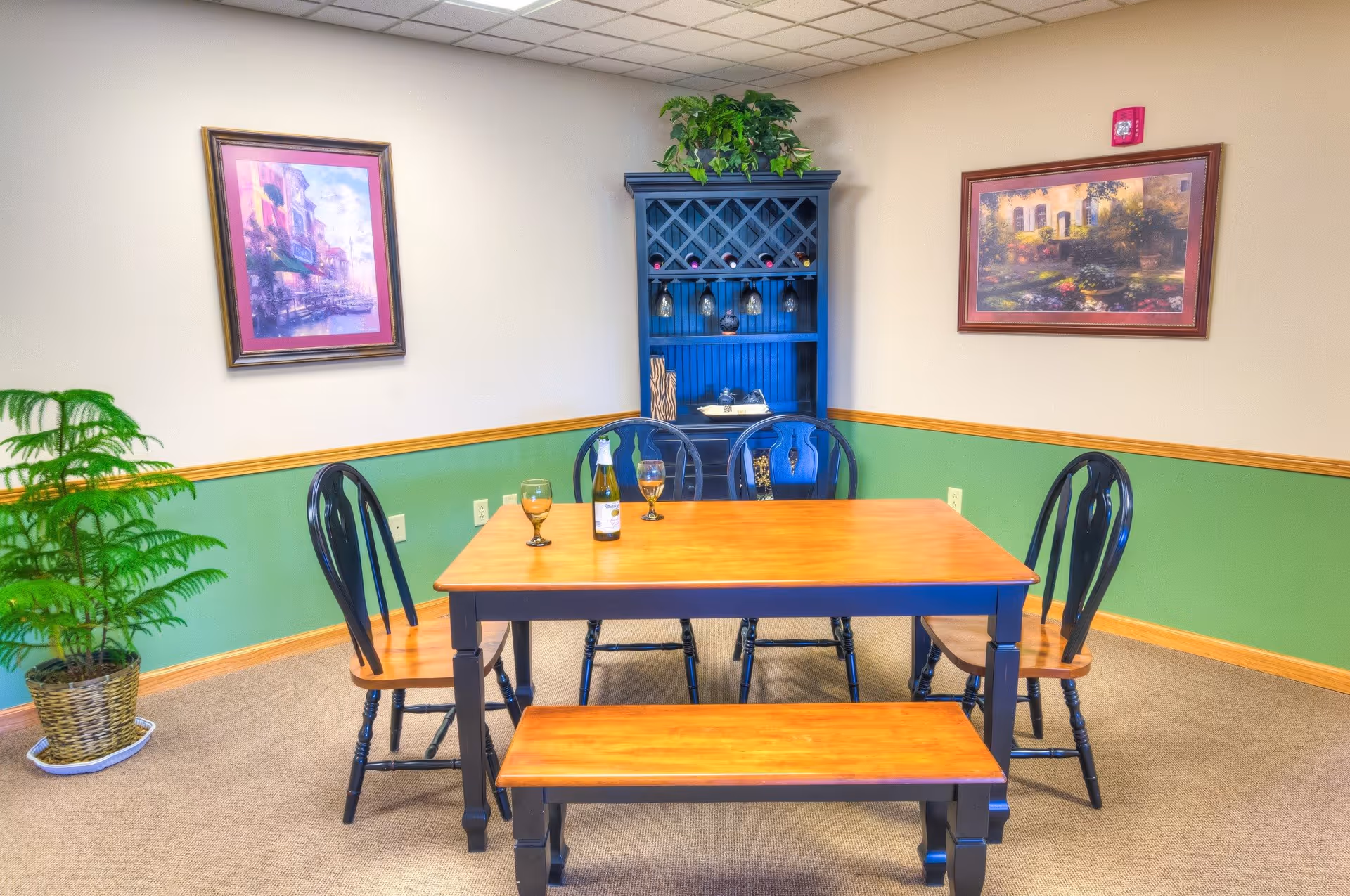 A dining area with a wooden table, four chairs, and a bench. On the table are two wine glasses and a bottle of wine. Behind the table is a blue cabinet with wine storage and hanging wine glasses. The walls are painted beige and green with wooden trim, and there are two framed paintings on the walls. A potted plant is placed in the corner of the room.