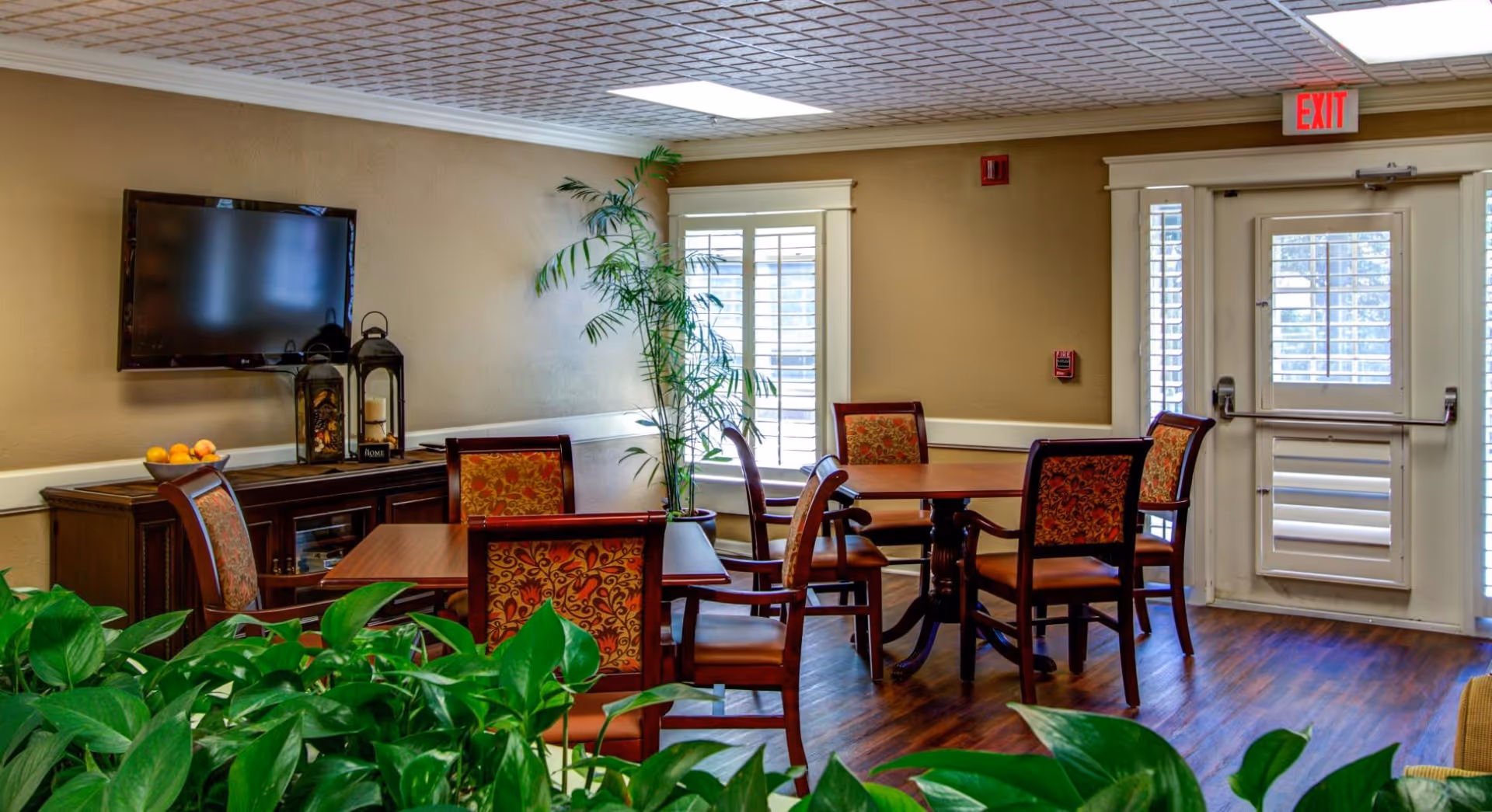 A cozy common area with multiple wooden tables and chairs featuring patterned upholstery. There is a flat-screen TV mounted on the wall above a wooden cabinet with decorative lanterns and a bowl of fruit. Large green plants are in the foreground, and windows with white shutters and a door with an exit sign are visible in the background.