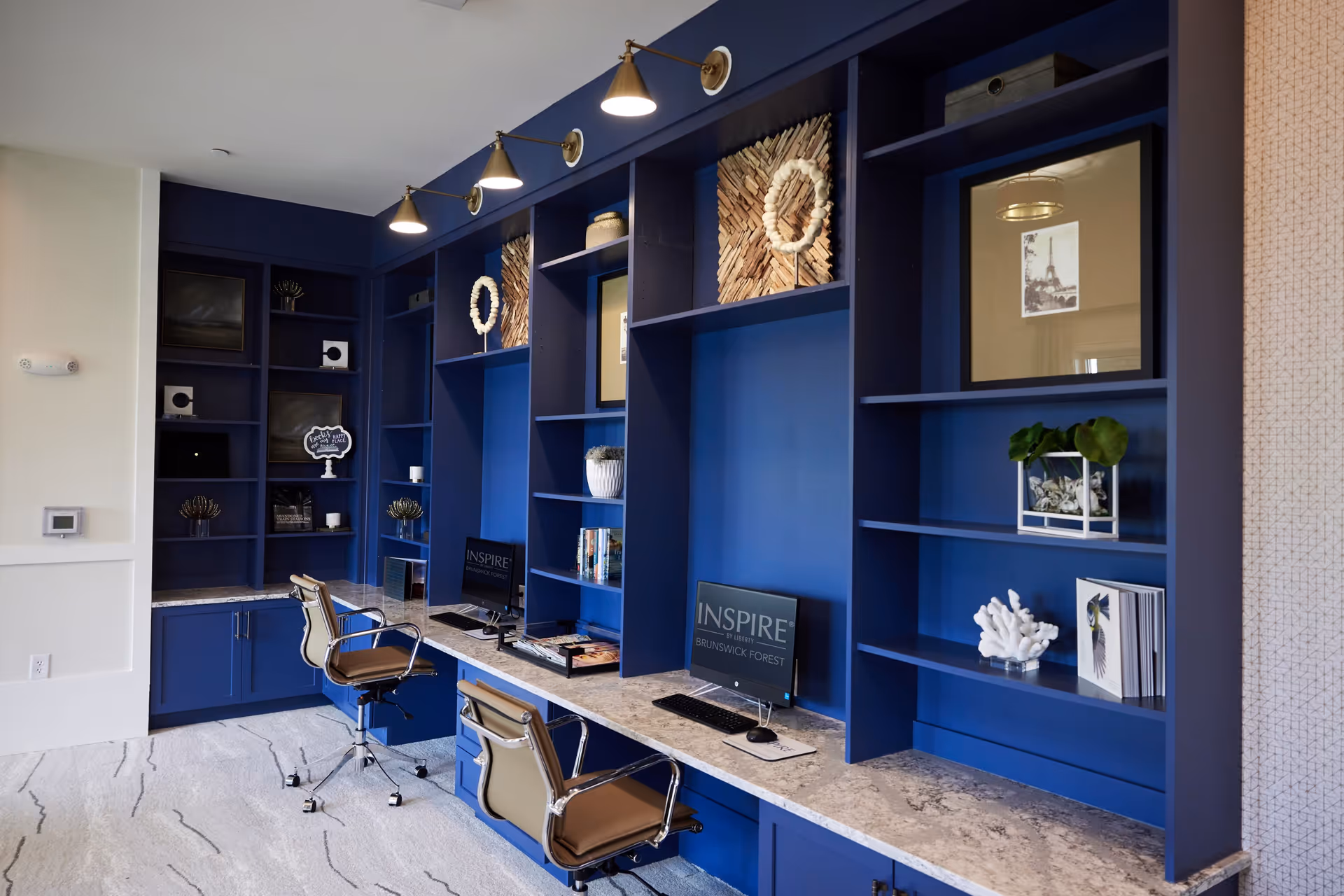 A modern workspace area with blue built-in shelves and a marble countertop. Two office chairs with brown seats and chrome frames are positioned in front of two computer monitors displaying the text 'INSPIRE BRUNSWICK FOREST'. The shelves are decorated with various art pieces, plants, and decorative objects. The room has a light-colored carpet with a subtle pattern and wall-mounted lights above the shelves.
